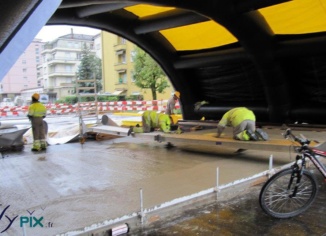 Tente de chantier gonflable: des ouvriers travaillent la pose de béton au sol, en étant protégé de la pluie et des intempéries. Les tentes, abris et hangars gonflables en enveloppe PVC sont de plus en plus utilisés dans le BTP. Ils présentent des avantages considérables pour les professionnels du secteur en termes de sécurité, de temps de montage et de démontage, ainsi que de protection contre les intempéries. – Droits réservés, reproduction interdite. Copyright FlyPix / Vivien LAÏLLE.
