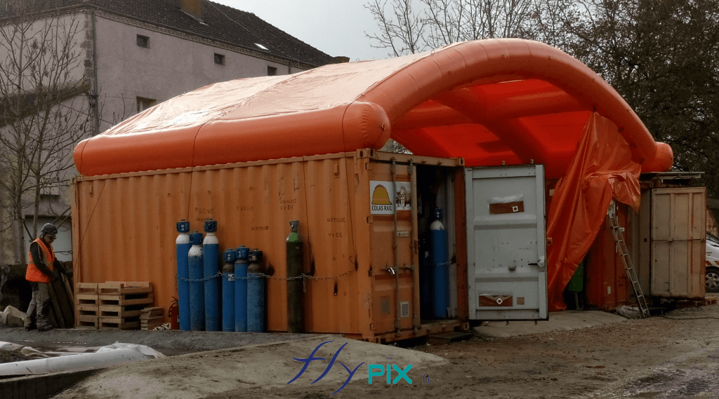 Un préau gonflable en forme de demi-lune, entre 2 containers maritimes.