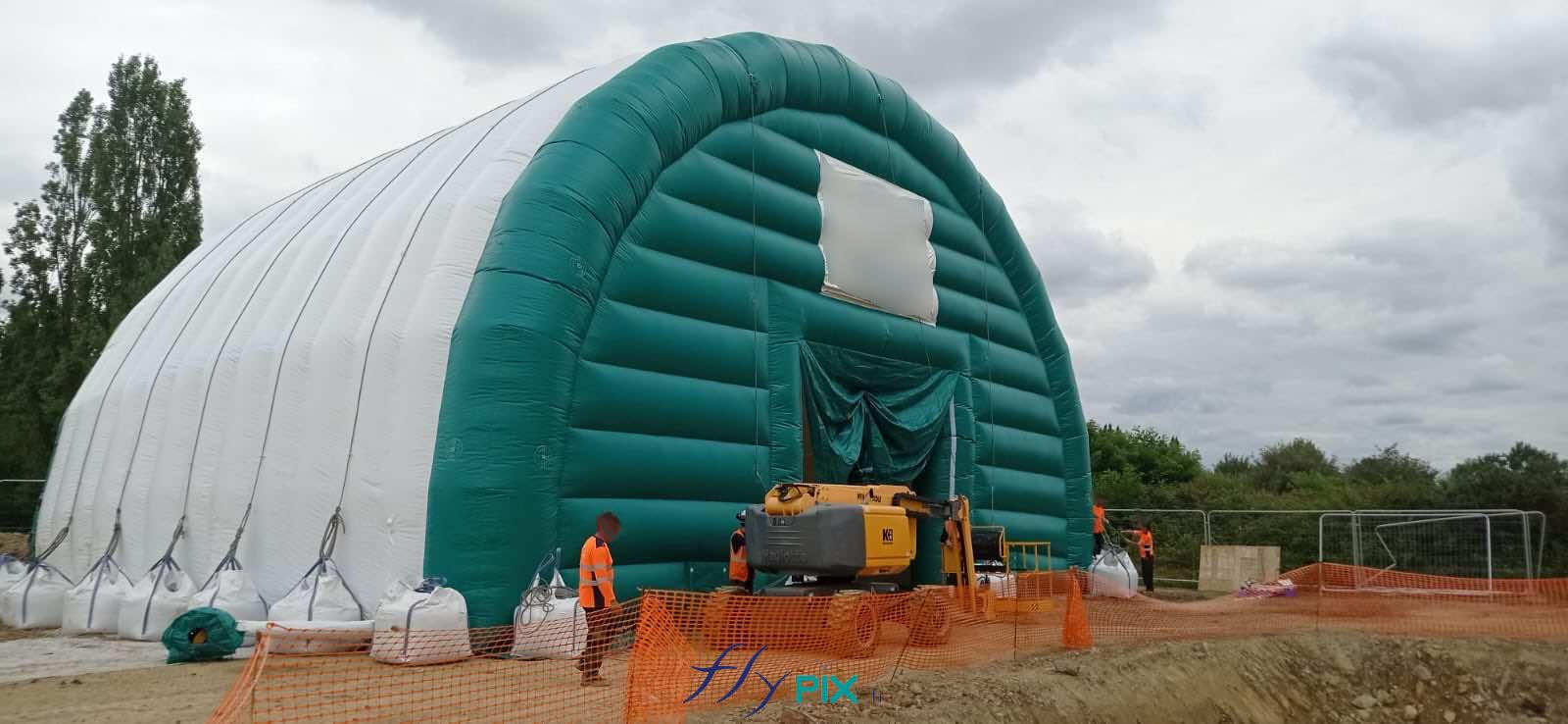 , Chantier de dépollution de site : 2 modules de hangar gonflable de S = 200 m2, soit S = 400 m2 de surface utile de travail,  Une photographie de la face du pignon avant de la tente gonflable de chantier ; le pignon est capitonné en enveloppe PVC 0.45 mm double peau, une grande porte d’accès en enveloppe PVC simple peau 0.45 mm permet l’accès aux machines imposantes de grandes tailles. A gauche et à drotie de la porte principale, en bas, vous pouvez voir les 2 emplacements prévus pour les manchons à air pour le contrôle du climats et la filtration des poussières contaminées en provenance des sols à l’intérieur. La taille des techniciens sert d’echelle pour démontrer l’aspect imposant du hangar gonflable de chantier. – Photo : copyright FlyPix/Vivien LAÏLLE, droits réservés.