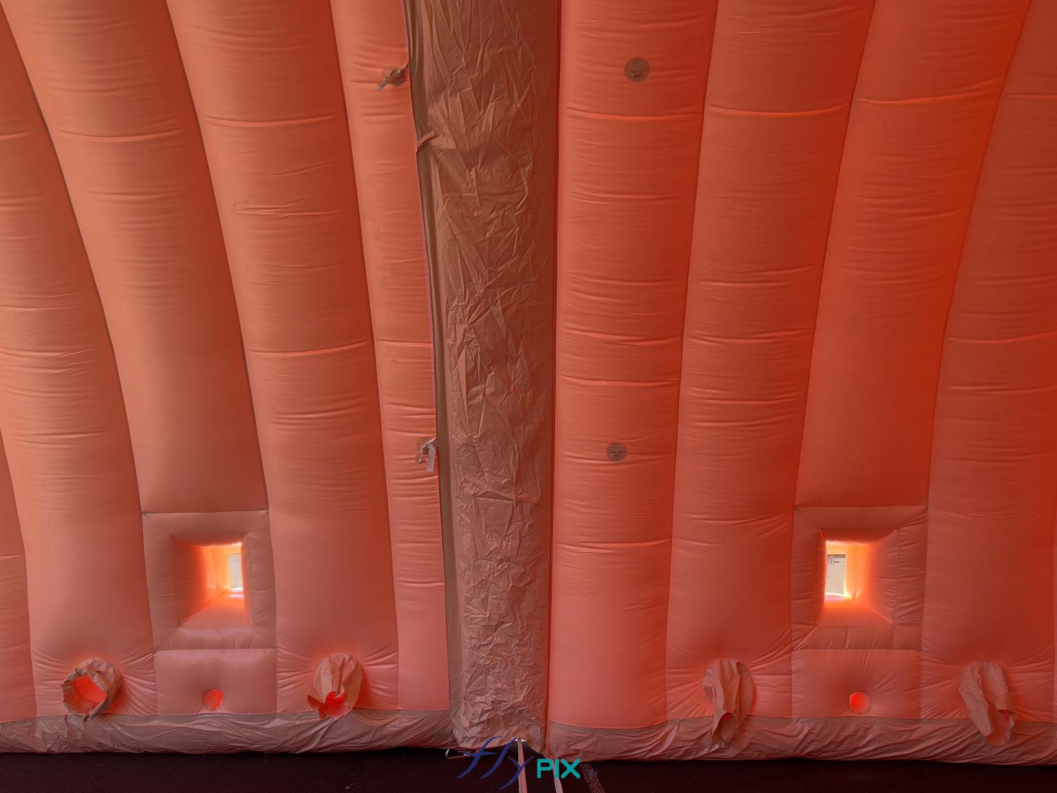 , Hangar gonflable militaire L = 25 m x l = 14 m x H = 7 m, S = 350 m2,  Sur cette photo, 2 modules sont assemblés et jointés, avec les fenêtres transparentes et les volets pliables et amovibles, qui permettent de disposer d'une source de lumière naturelle à l'intérieur d'un module de hangar gonflable. Dans les parties inférieures des modules gonflables, nous voyons les manchons à air et les trous passe fils et gaines électriques. - Copyright : Vivien LAÏLLE/FLYPIX, droits réservés.