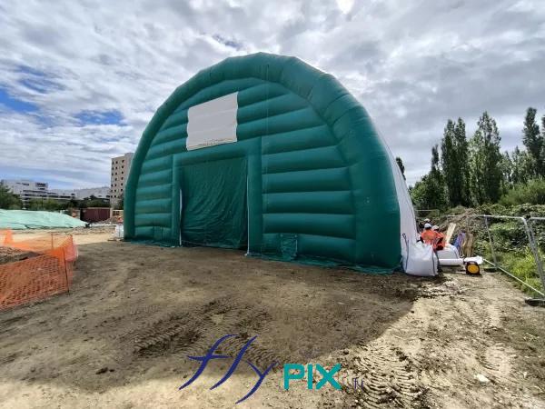 Vue extérieure depuis la façade avant, le pignon avant d'un module de grand hangar gonflable de chantier de dépollution de site, avec la présence d'une grande porte principale pour l'accès des engins et des machines de BTP. Abri en enveloppe PVC 0.45 mm double peau capitonnée, ventilé par turbines en permanence. - Photo : copyright FlyPix/Vivien LAÏLLE, droits réservés. Vue extérieure depuis la façade avant, le pignon avant d'un module de grand hangar gonflable de chantier de dépollution de site, avec la présence d'une grande porte principale pour l'accès des engins et des machines de BTP. Abri en enveloppe PVC 0.45 mm double peau capitonnée, ventilé par turbines en permanence. - Photo : copyright FlyPix/Vivien LAÏLLE, droits réservés.
