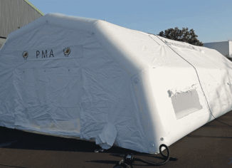 Une vue générale de l'abri gonflable poste de premiers secours, de dimensions L = 10 m x l = 6 m, soit une surface utile intérieure de S = 60 m2. Sur cette photo, nous voyons la façade du pignon arrière de la tente gonflable PMA, avec une grande porte de sortie. Sur les murs de la toiture, nous observons des fenêtres et des points de jour, avec moustiquaires, PVC transparent et volets amovibles. C'est une tente air captif, c’est-à-dire que les boudins d'ossature en enveloppe PVC 0.6 mm simple peau sont étanches, ils sont gonflés avec une pompe électrique via une valve anti-retour et un régulateur de pression. A gauche et à droite de la porte, il y a 1 manchon à air.