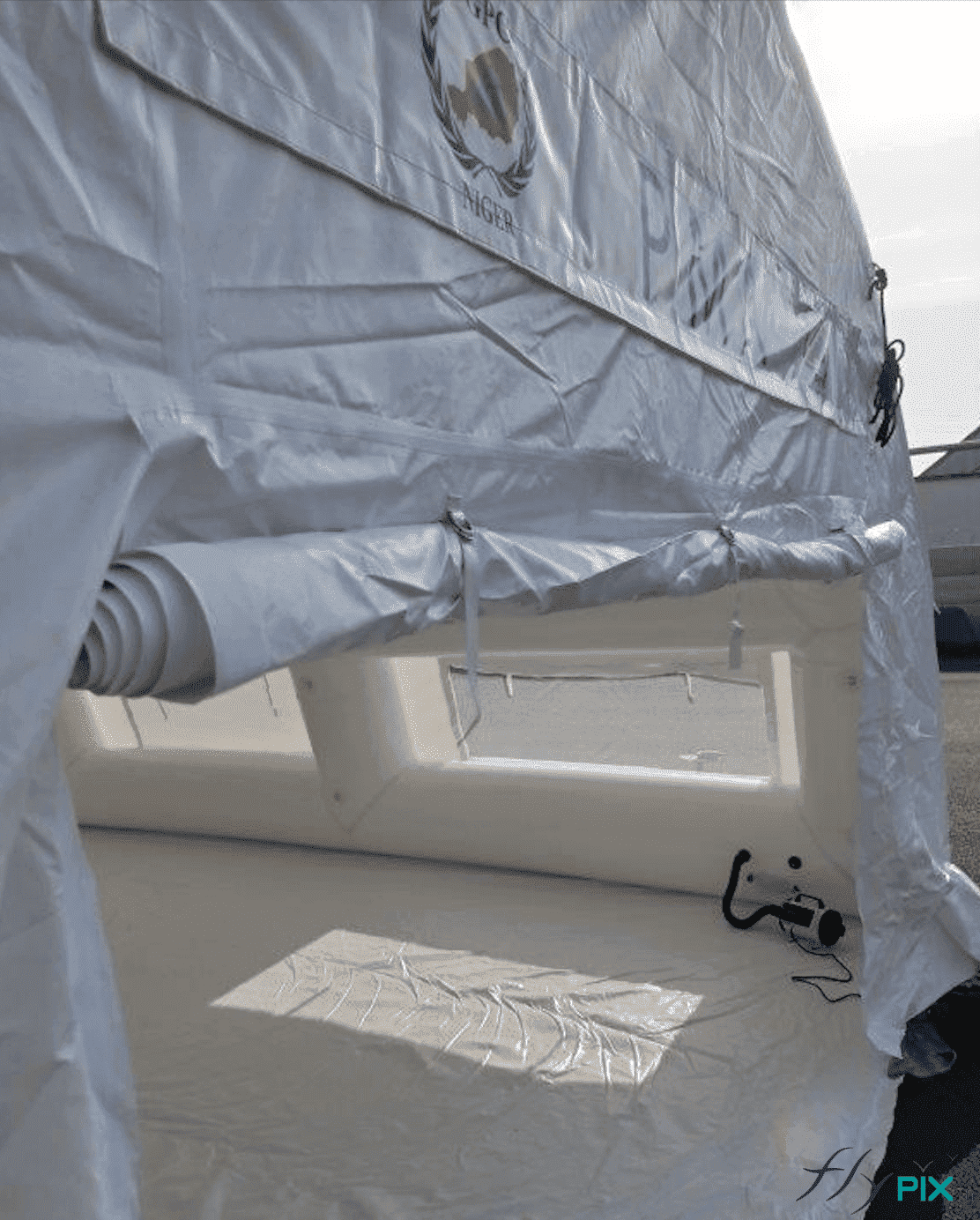 Une autre vue de la grande porte de sortie, sur le pignon arrière de la tente gonflable PMA. La porte est pliée et la bâche au sol déployée. Au fon, on peut voir des valves et un régulateur de pression en fonctionnement. Une autre vue de la grande porte de sortie, sur le pignon arrière de la tente gonflable PMA. La porte est pliée et la bâche au sol déployée. Au fon, on peut voir des valves et un régulateur de pression en fonctionnement.