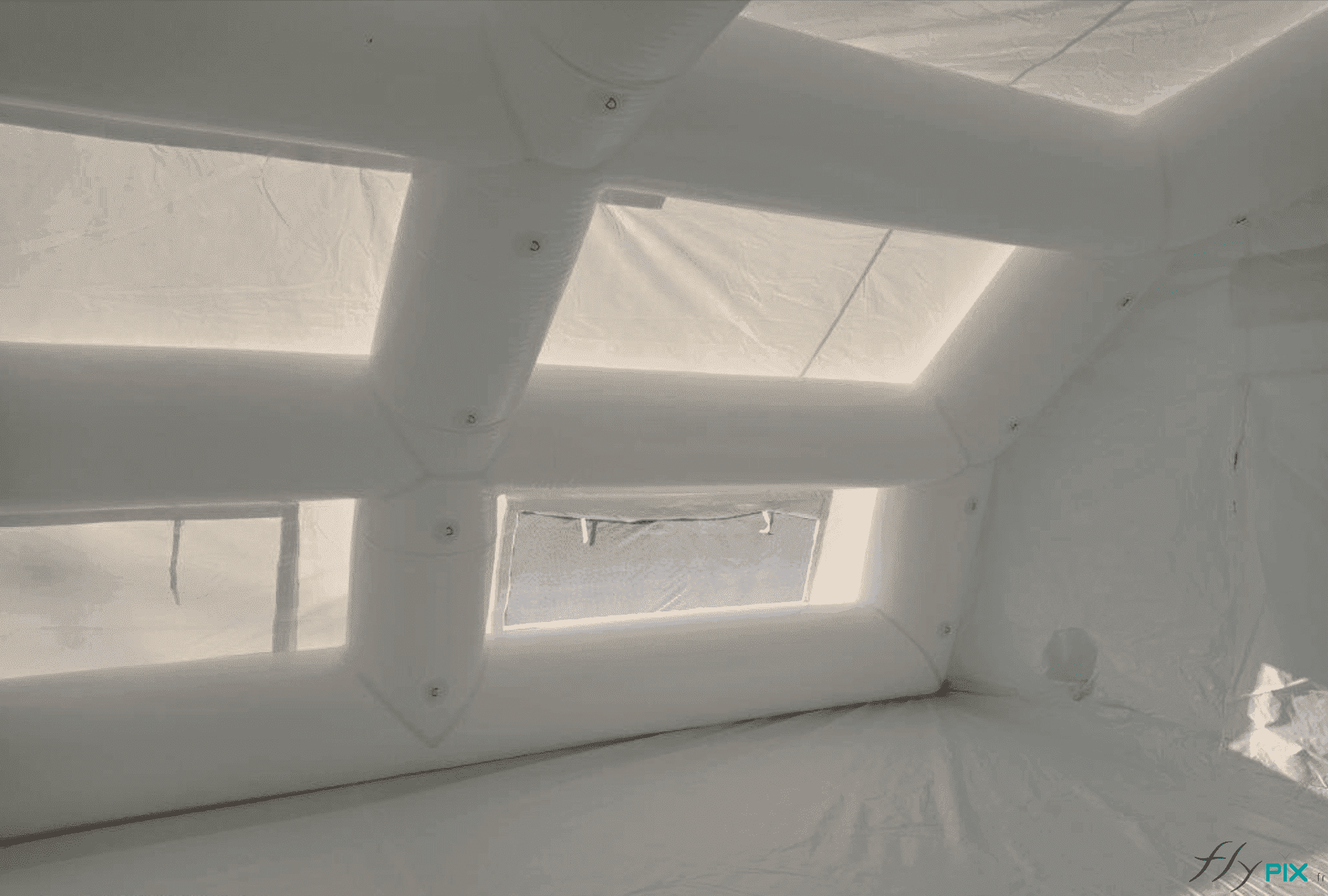 Une vue de l'intérieur de la tente gonflable PMA de surface S = 60 m2, avec une bâche au sol en PVC, amovible. Les boudins d'ossature, squelette de l'abri gonflable procurent une très grande solidité et robustesse à la structure gonflable. Ils sont tous interconnectés et ils présentent, à des espacements réguliers, des anneaux et des œillets métalliques de fixation, très utiles pour y attacher des accessoires médicaux, ou des dispositifs éclairages LED. Notez que l'enveloppe en PVC de couleur blanche et la présence de points de jour et de fenêtres permettent d'obtenir une lumière intérieure naturelle et diffuse. Une photo qui montre les détails et les finitions des boudins d'ossatures et des fenêtres faisant office de point de jour pour éclairer l'intérieur de la tente gonflable PMA d'une lumière naturelle et diffuse. Une bâche au sol, en enveloppe PVC lavable, est amovible. Elle a l'avantage de permettre aux médecins, docteurs et infirmiers de disposer d'un environnement intérieur sain, à l'abri de l'humidité, et avec une température stable ou un climat / atmosphère contrôlé. Sur les ossatures interconnectées, il y a aussi des anneaux et des œillets de fixation métalliques qui sont des interfaces utiles utiles et ergonomiques pour y attacher des accessoires médicaux ou des éclairages LED.
