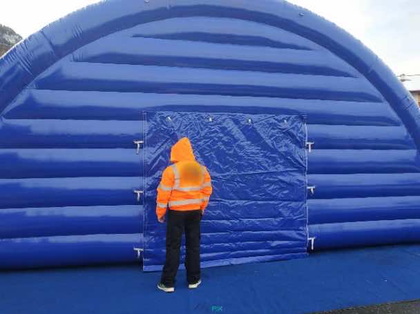 Cette photo montre une vue extérieure du pignon avant, avec la porte d'accès et le mur en enveloppe PVC 0.6 mm double peau capitonnée. La personne sur la photo permet de disposer d'une échelle de référence pour la hauteur et les dimensions. Ce hangar gonflable est imposant. Cette photo montre une vue extérieure du pignon avant, avec la porte d'accès et le mur en enveloppe PVC 0.6 mm double peau capitonnée. La personne sur la photo permet de disposer d'une échelle de référence pour la hauteur et les dimensions. Ce hangar gonflable est imposant.