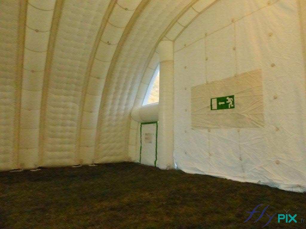 Sur cette vue intérieure d'un abri gonflable, nous voyons sur le pignon avant d'un module, une grande porte d'accès principale et une porte de service secondaire plus petite. Notez la présence de signalétiques d'issues de secours, obligatoires pour les tentes gonflables de réception de public. Sur cette vue intérieure d'un abri gonflable, nous voyons sur le pignon avant d'un module, une grande porte d'accès principale et une porte de service secondaire plus petite. Notez la présence de signalétiques d'issues de secours, obligatoires pour les tentes gonflables de réception de public.