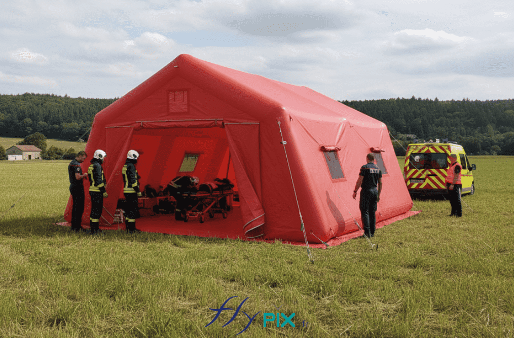 Une vue de simulation montrant le déploiement d'une tente PMA de surface 37 m2, une structure gonflable air captif, gonflée à l'air avec une pompe électrique. Un déploiement en zone de campagne, en zone rurale. Cet abri gonflable est en enveloppe PVC 0.6 mm simple peau pour les ossatures de boudins gonflable étanches, et en enveloppe PVC 0.45 mm simple peau pour les murs et la toiture. Cette tente gonflable est une structure facile à installer et déployer, et une fois pliée elle prend très peu de place dans le véhicule d'intervention. Il y a une bâche au sol amovible qui permet de dsposer d'un sol sain et propre. - Cette image a été générée par IA, grâce à GEMINI, à partir des vues de modélisations en 3D du BAT (bon à tirer) créées par notre infographiste et validées par le commanditaire, et aussi à partir de photos et de prises de vues réelles de certains détails de la tente gonflable, prises directement dans notre atelier de fabrication. Média partiellement généré par une IA et vérifié par un agent. - Copyright Vivien LAÏLLE/FlyPix, droits réservés, reproduction interdite.