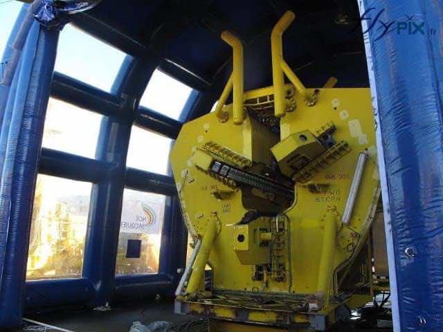 Une vue extérieur de la façade du pignon avant du hangar gonflable en forme de U-inversé, avec une très grande et haute porte permettant de faire rentrer à l'intérieur un gros moteur de bateau, dans le cadre d'un chantier industriel naval, dan sun port de navires.