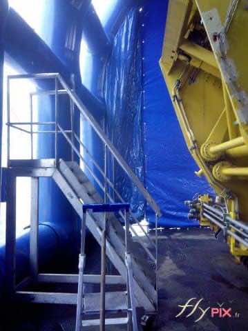 Une vue intérieure d'un hangar gonflable de chantier industriel, pour un chantier naval : murs en enveloppe PVC et grandes fenêtres et points de jour en PVC Crystal transparent, pour laisser passer la lumière naturelle. Il s'agit ici d'une tente gonflable de confinement, pour le nettoyage et la maintenance d'un gros moteur de bâteau. Pour empêcher la dissémination de produits chimiques ou de poussières poulluantes vers l'environnement extérieur, et pour la protection de la zone de travail contre les intempéries.