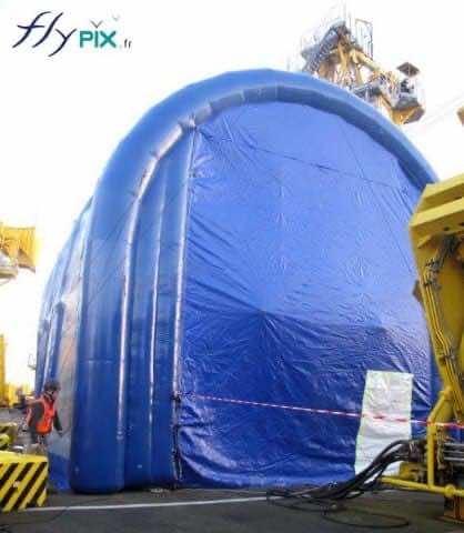 Hangar gonflable TECHNIP : vue extérieur de la façade du pignon avant, avec une très grande porte, ici fermée, lors d'un chantier naval pour la maintenance et le nettoyage dun gros moteur d'un navire. Ce hangar gonflable permet de travailler à l'abri des intempéries et d'assurer un confinement idéal.