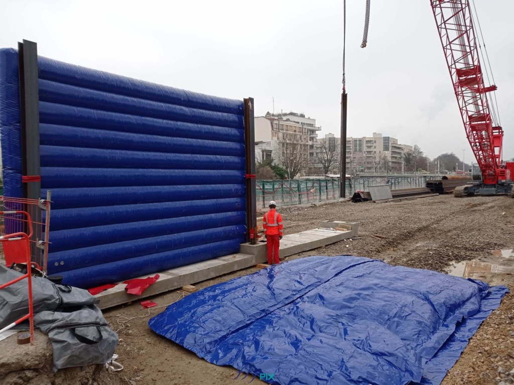 Grand Paris Express, chantier urbain de Meudon : cette photo est intéressante parce qu'elle montre plusieurs choses essentielles qui concerne les structures gonflable de murs de réduction de bruits. Premièrement, au premier plan, au sol, un écran gon flable est déployé sur toute sa surface, dans l'attente de son gonflage à l'air avec une pompe électrique. C'est une structure air captif. Deuxièmement, en arrière plan, nous observons un mur gonflé, positionné verticalement sur une structure en béton et avec des poutres métalliques. Cette configuration procure une très grande résistance structurelle aux vents, tout en minimisant au maximum l'emprise au sol, en limitant la présence et l'utilisation de sacs de lestages à remplir de sable. Tout en conservant une grande mobilité et un très bon réemploi. Grand Paris Express, chantier urbain de Meudon : cette photo est intéressante parce qu'elle montre plusieurs choses essentielles qui concerne les structures gonflable de murs de réduction de bruits. Premièrement, au premier plan, au sol, un écran gon flable est déployé sur toute sa surface, dans l'attente de son gonflage à l'air avec une pompe électrique. C'est une structure air captif. Deuxièmement, en arrière plan, nous observons un mur gonflé, positionné verticalement sur une structure en béton et avec des poutres métalliques. Cette configuration procure une très grande résistance structurelle aux vents, tout en minimisant au maximum l'emprise au sol, en limitant la présence et l'utilisation de sacs de lestages à remplir de sable. Tout en conservant une grande mobilité et un très bon réemploi.